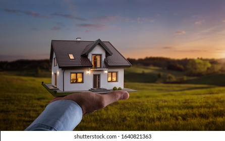 Concept Of Buying Or Building New Home. Male Hand Showing, Offering A New Dream House At The Empty Field With Copy Space