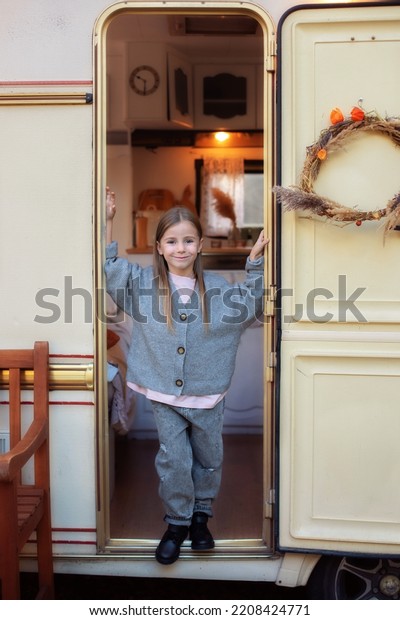 Concept camping, outdoor, nature, adventure. Smiling\
little girl in casual clothes standing on porch RV house in garden.\
Cute young girl stand near trailer door. Child in cozy campsite\
fall backyard. 