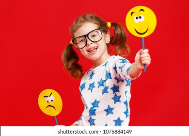 Concept Of Children's Emotions. Child Girl Chooses Between A Sad And Joyful Smile On  Colored Red Background
