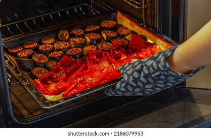 The concept of cooking, cooking. A woman with a potholder takes out a baking dish with vegetables from the oven in a home kitchen. The housewife is cooking dinner. Baked peppers and eggplants. Food. 