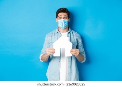 Concept of coronavirus, quarantine and social distancing. Young man searching apartment, showing house paper model, wearing medical mask, renting or buying propery, blue background