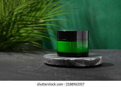 Concept of cosmetic face cream products in glass jar with natural moisturizer ingredients on black marble podium on green background decorated fresh leaves with shadow. Close up.