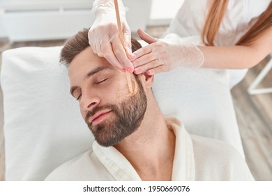 Concept Of Cosmetology And Facial. A Woman Beautician Makes Face And Beard Modeling For A Man Waxing Epilation. Depilation With Wax