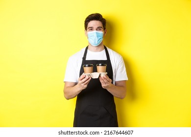 Concept Of Covid-19, Cafe And Social Distancing. Barista In Medical Mask Serving Coffee In Takeaway Cups, Standing In Black Apron Against Yellow Background