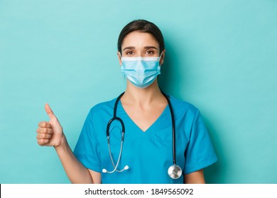Concept of covid-19 and quarantine concept. Close-up of confident female doctor in medical mask and scrubs, holding stethoscope, showing thumbs-up, standing over blue background