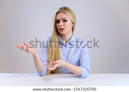Concept cute model talking to the camera sitting at the table. Close-up portrait of a beautiful blonde girl with excellent makeup with long smooth hair on a white background in a blue shirt.