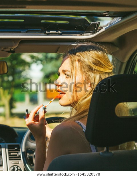 Concept of danger\
auto drive. Young woman driver painting her lips doing applying\
make up while driving the\
car.