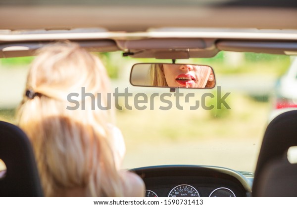 Concept of danger\
auto drive. Young woman driver painting her lips doing applying\
make up while driving the\
car.