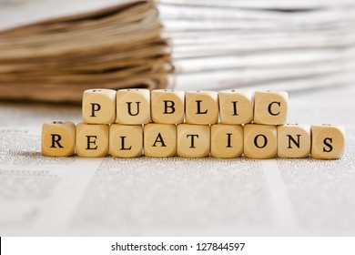 Concept of dices with letters forming words: Public Relations. Generic newspaper background with some blurred text on the bottom and paper stack in the back. - Shutterstock ID 127844597