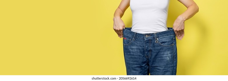 The Concept Of Diet, Proper Nutrition, Weight Loss. Slim Woman Showing Loose Jeans And Her Loss Weight. Woman White T-shirt And Oversize Jeans Isolated On Yellow Background. Banner With Copy Space.