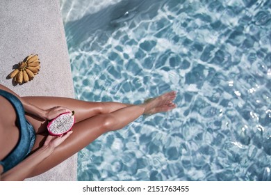 Concept of diet, spa and cosmetics. Enjoying tropical fruits, suntan and vacation. Young slim woman in green swimsuit sitting near swimming pool holding dragon fruit.