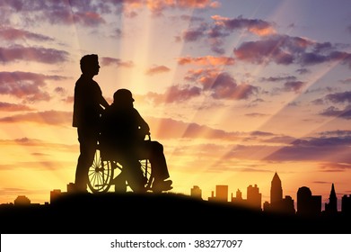 Concept of disability and old age. Silhouette of a man looking after a disabled person on a background of city sunset