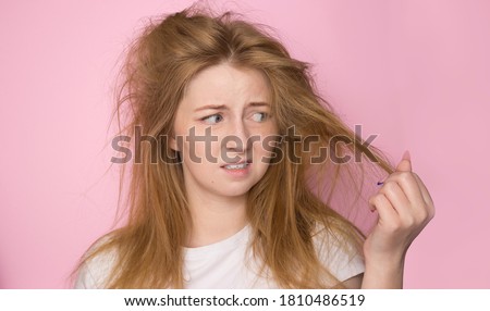 The concept of dry lifeless hair. A woman on a pink background holds her disheveled, tangled hair and looks at it with a discontented expression.