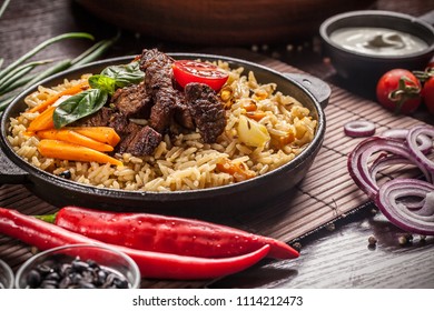 The Concept Of Eastern, Uzbek Cuisine. Pilaf, Plov In A Black Cast-iron Frying Pan On A Wooden Table, Next To Lie Ingredients, Onions, Tomatoes, Parsley, Pepper. Central Asian Cuisine.