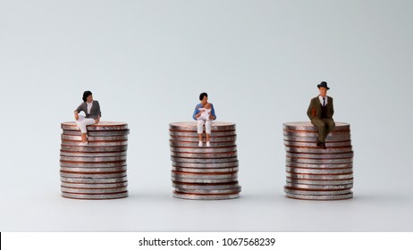 The Concept Of Equal Pay. Miniature women holding baby sitting on a pile of coins and two miniature people sitting on a pile of coins.