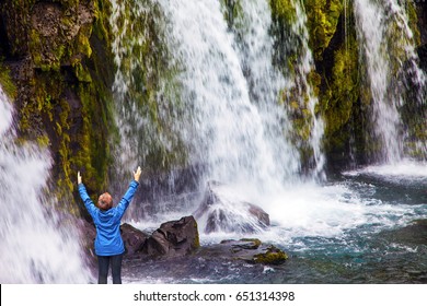 Concept Of Exotic And Extreme Tourism. Waterfall Kirkjoufellfoss. Elderly Woman -  Traveler Enthusiastically Raised Her Hands. Summer In Iceland