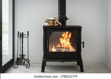 Concepto de chimenea con fuego en casa. Confortable sala de estar con madera de tronco encima de la chimenea de metal, detrás de puerta de cristal y diseño interior moderno