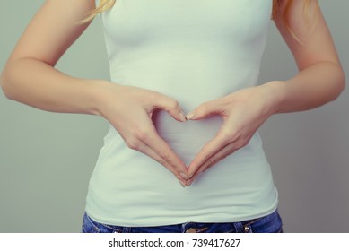 Concept Of First Weeks Pregnancy. Woman's Hands Forming Heart On Her Tummy. Concept Of Healthy Eating And Lifestyle. Woman In White T-shirt And Jeans Isolated Against Grey Background