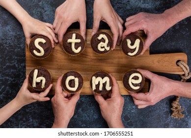 Concept flat lay Stay Home. Many hands of different ages of people reach out and hold chocolate muffins, with cream letters. Eight cupcakes make up the two words. Big family on self-isolation.