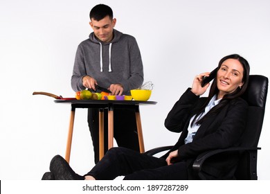 The Concept Of Gender Stereotypes. A Man Is Ready In The Kitchen And A Business Woman Talks On The Phone.