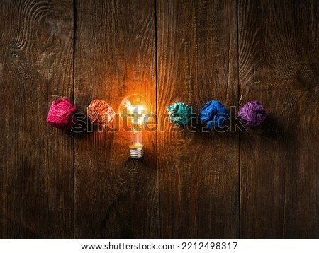 Concept of a great creative idea with crumpled colorful paper and a light bulb emitting a luminous flux on a textured wooden table.