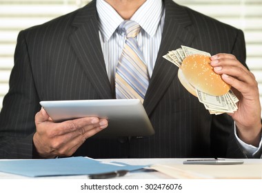 Concept For Greedy Corporate Executives, Businessman Is Eating A Burger Stuffed With Money And Browsing On A Tablet Computer In His Office