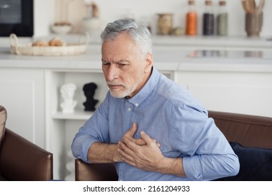Concept of health and cardiovascular problems. Senior man with cardiology disease feeling sharp heart pain, holding hands on chest, sitting on armchair at home. Pensioner need emergency medical care