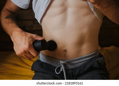 The Concept Of Healthy Lifestyle. Caucasian Man Gives Himself Massage With Percussion Gun Inside. Athlete Kneads Abdominal Muscles And Abs After Training While Sitting On Bed.