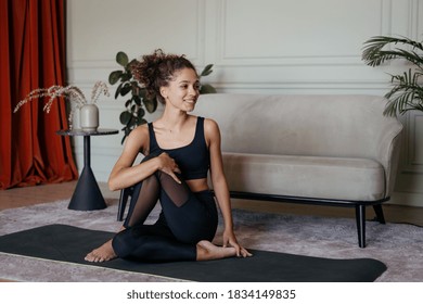 Concept of healthy lifestyle. Happy and adult african american woman training at home, sitting on sport mat, making exercise, smiling wide and stretching upper body