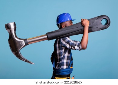Concept of heavy work, African american builder with giant hammer in studio shot. Strong man heavy work Construction worker carrying huge work tool wearing coveralls and hard hat, construction