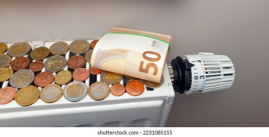 The concept of the high cost of heating and electricity and the impending energy crisis. On a heating radiator with a thermostat, a pack of 50 euro banknotes and a lot of coins - Shutterstock ID 2231085155