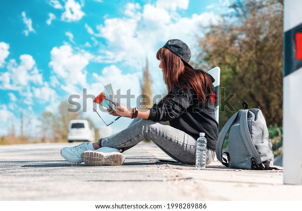 Concept of hitchhiking and local travel. A hipster\
woman in cap with tattoed hands sitting along the road and reading\
a paper map. Copy\
space.