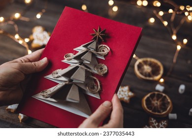 Concept of holiday handicraft at home, family quality time with children. Woman holding handmade craft paper Christmas New Year card with anise star. Lights, gingerbread cookies on wooden background - Powered by Shutterstock