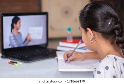 Concept of homeschooling or e-learning, young girl busy in writing by looking into laptop while teacher explaining during covid-19 or coronavirus pandemic crisis