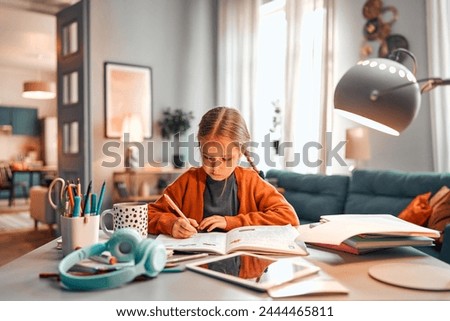 Concept of homework. Diligent elementary school student sitting by table with educational materials at living room. Concentrated girl making exercises at workbook and writing in answers with pen.