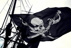 Concept Of Illegality And Piracy: Detail Of A Real Pirate Flag With Skull And Shinbone Called Jolly Roger Fluttering In The Wind Among The Rigging Of An Ancient Wooden Pirate Ship