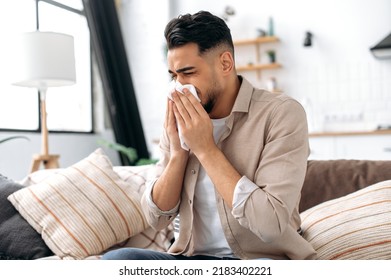 The concept of illness, runny nose. Unhealthy sad arabian or indian man sits at home in the living room on the couch, feels unwell, suffers from a runny nose, sneezing, uses napkins, needs treatment - Powered by Shutterstock