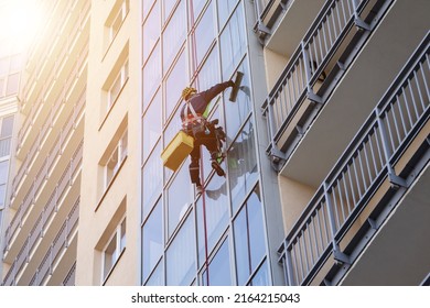 Concept of industry urban works. Industrial mountaineering worker in uniform hangs over residential facade building, washing exterior glazing. Rope access laborer hangs on wall of house. Copy space - Shutterstock ID 2164215043