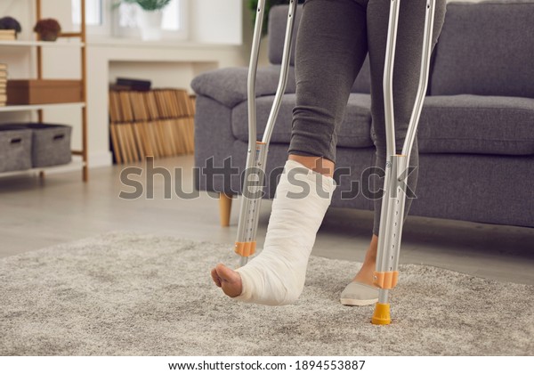 Concept of injury in domestic or car accident,\
rehabilitation, making progress and successful recovery. Young\
woman with broken leg in plaster cast stands up from couch and\
walks with crutches at\
home