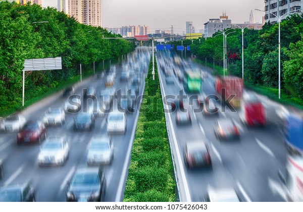 Concept of interaction between nature and\
technology - green trees and dividing green line along busy road,\
protect environment. Cars on highway in busy city, Air pollution.\
Photo with blur in\
motion.