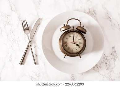 concept of intermittent fasting, ketogenic diet, weight loss. fork and knife crossed and alarmclock on a plate 