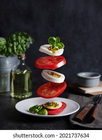 Concept Levitation Food. Italian Caprese Salad With Tomatoes, Mozzarella Cheese, Basil And Pesto Sauce Fly Over The Plate On A Dark Background. Front View Image