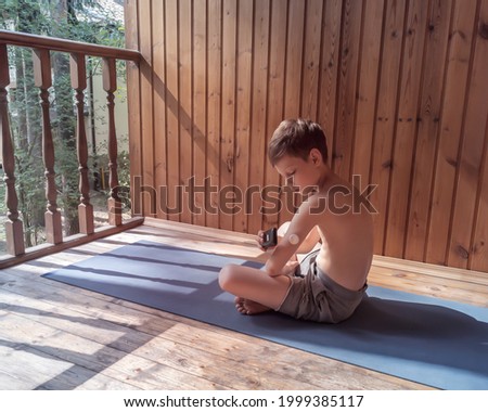 Concept of life of a child and sport with diabetes and glycemic control. Summer vacation: a boy sitting on the gym mat in the open air and measuring sugar levels using a sugar monitoring system