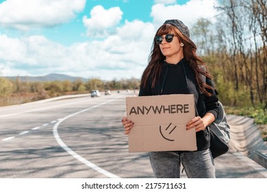 The concept of local traveling and hitchhiking. Portrait of smiling young woman in sunglasses and cap holding a cardboard sign with text anywhere.