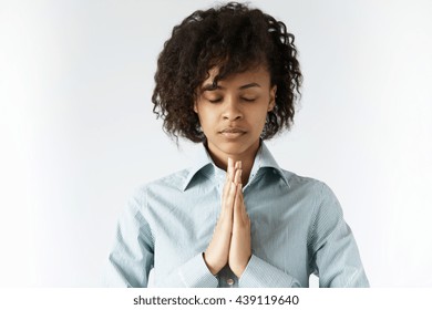 Concept of meditation and prayer. African female standing in meditative pose against white wall, with closed eyes and calm face full of sense of inner peace and wisdom, holding hands in prayer
