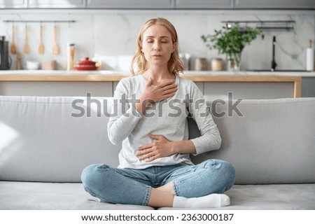 Concept of mental health. Woman sitting on couch and doing calming breathing exercises after panic attack. Female inhaling and exhaling to deep breath. Self-control, anxiety relief concept