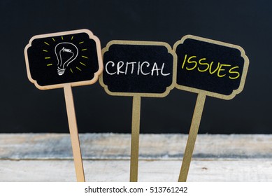 Concept Message CRITICAL ISSUES And Light Bulb As Symbol For Idea Written With Chalk On Wooden Mini Blackboard Labels, Defocused Chalkboard And Wood Table In Background