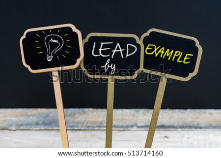 Concept message LEAD BY EXAMPLE and light bulb as symbol for idea written with chalk on wooden mini blackboard labels, defocused chalkboard and wood table in background