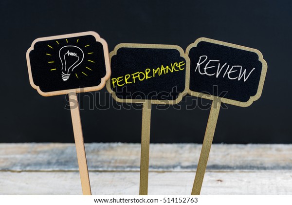Concept message\
PERFORMANCE REVIEW and light bulb as symbol for idea written with\
chalk on wooden mini blackboard labels, defocused chalkboard and\
wood table in\
background