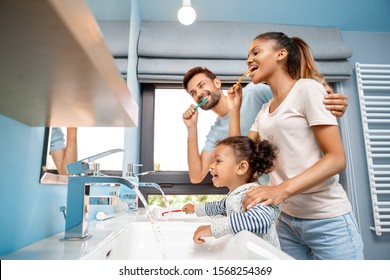 Concept of mixed race family. Low angle view of cheerful young adult man hugging happy woman and brushing teeth at morning. Father and mother standing together with african daughter in bathroom - Shutterstock ID 1568254369